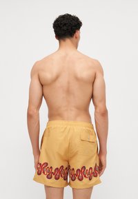 Gelbe Schwimmshorts mit einem gewellten rot-schwarzen Muster am Saum, elastischem Bund und einer hinteren Tasche mit Druckknopfverschluss.