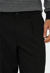 Schwarze Hose mit glatter Textur, ausgestattet mit einem einzelnen Knopfverschluss, gefälteter Vorderseite und seitlichen Taschen, präsentiert vor einem gemusterten Oberteil.