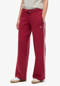 Pantalones de chándal burdeos de tela suave con cintura ajustable, bolsillos laterales y rayas blancas en contraste a lo largo de los lados. Corte relajado.