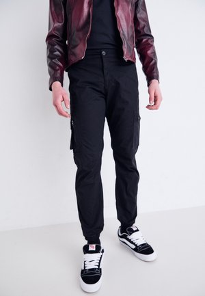 Person iført sorte cargobukser, sorte og hvide sneakers, sort skjorte og en mørkerød læderjakke, der står mod en hvid baggrund.