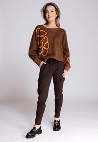 Brauner Strickpullover mit orangefarbener Blumenstickerei, kombiniert mit dunkler Cargo-Hose und schwarzen Loafers mit grober Sohle.