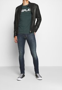 Man wearing black leather jacket, dark green t-shirt with white text, dark blue jeans, and black high-top sneakers with white laces.