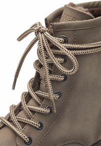 Botín de cuero marrón hasta el tobillo con superficie texturizada, puntera redonda, ojales negros para los cordones y cordones gruesos de color marrón trenzados. Forro de forro polar visible en la parte superior.