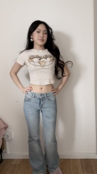 Ung kvinde med langt, mørkt hår iført en cropped "Baby Girl" t-shirt og lyseblå jeans, står med hænderne på hofterne mod en hvid væg.
