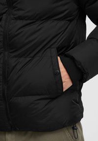 Black puffer jacket featuring a zip closure, ribbed cuffs, and a side pocket. The material appears smooth with a slightly glossy finish.