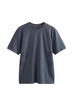 Ung mand med tatoveringer iført en mørkegrå t-shirt, lysegrå shorts, grå sokker og sorte sneakers, stående mod en ensfarvet væg.