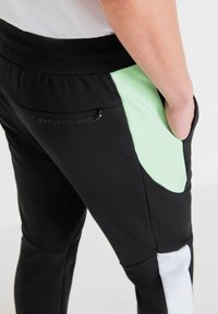Zwarte sportbroek met een mintgroene zijpaneel en een ritszak. Het logo "ANTONYMORATO" is in het zwart gedrukt op de achterzak.