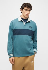 Νέος άνδρας με κατσαρά μαλλιά φορώντας μπλε-πράσινο μακρυμάνικο πουκάμισο ράγκμπι με λευκό γιακά και μπεζ παντελόνι φόρμας New Balance, στέκεται μπροστά σε απλό φόντο.