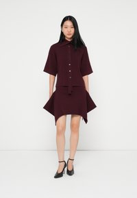 Vestido de malha canelada bordeaux com mangas curtas, gola, botões na frente e bainha assimétrica, combinado com sapatos de salto alto pretos.