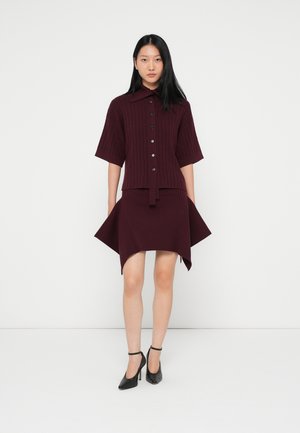 Vestido de malha canelada bordeaux com mangas curtas, gola, botões na frente e bainha assimétrica, combinado com sapatos de salto alto pretos.