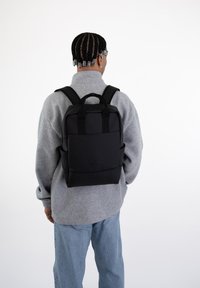 Mochila negra con un diseño estructurado, hecha de material suave, con correas de hombro acolchadas y un asa en la parte superior, sobre un fondo claro.