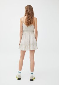 Vestido a rayas color beis claro con un corpinho ajustado y un dobladillo acampanado, que presenta tirantes finos y un borde con volantes. Combinado con zapatillas grises y amarillas.
