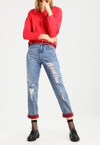 Rode sweatshirt met lange mouwen, gecombineerd met lichtblauwe, versleten wijde jeans met een rode geruite omslag en zwarte plataformen schoenen.