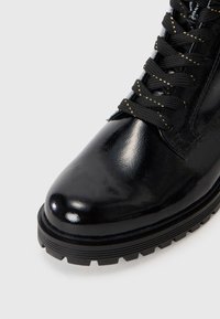 Botte en cuir verni noir avec une pointe arrondie, semelle en caoutchouc texturée et lacets noirs avec des accents dorés.