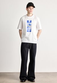 Λευκό βαμβακερό T-shirt με μπλε γραφική εκτύπωση με χαρακτήρες, συνδυασμένο με σκούρα τζην παντελόνια καμπάνα και μαύρα υποδήματα.