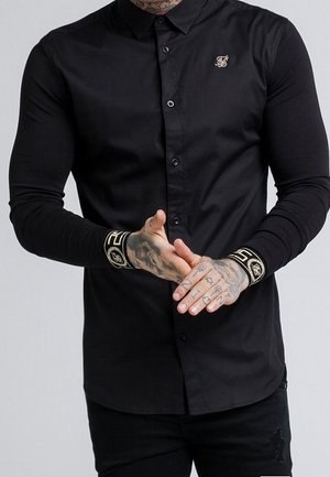 Mann trägt eng anliegendes schwarzes Hemd mit Knöpfen, mit goldbestickten Manschetten und kleinem goldenem Logo auf der Brust, Hände gefaltet und Tattoos sichtbar.