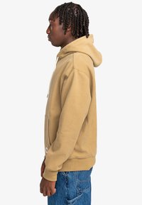 Beige hooded sweatshirt with ribbed texture, featuring a front pocket and drawstring. Worn with blue denim jeans. Profile view.