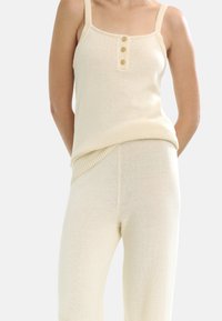 Jasno beżowy pleciony top na ramiączkach z cienkimi paskami, z guzikowym zapięciem. Połączony z pasującymi, lekkimi spodniami z dzianiny, tworząc spójną stylizację.