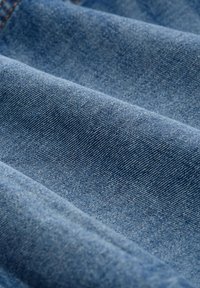 Denim tkanina u srednje plavoj nijansi s blago teksturiranom površinom, koja ima klasičnu tkaljsku strukturu i suptilne detalje u uzorku tkanja.