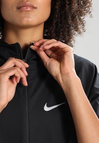 Černá zipová bunda s texturovanou tkaninou, vysokým límcem a bílým logem Nike na levé části hrudi. Ruce jsou umístěny blízko zipu.