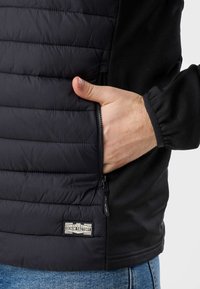 Hand with thumb out inserted into zippered pocket of black padded jacket with "Denim Factory" label, worn over blue jeans.