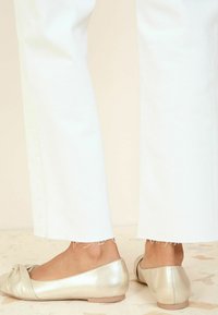 Metalen gouden ballet flats met een gedraaid stoffen accent, gecombineerd met witte wijde broekspijpen met rafelige zomen. Eenvoudig en strak ontwerp.