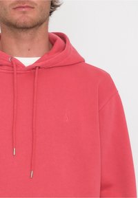 Sweat à capuche corail en tissu doux, doté d'une poche kangourou à l'avant, de cordons de serrage réglables et d'un petit logo de marque sur la poitrine.