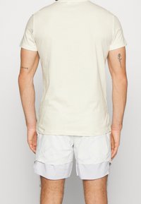 Ljusbeige t-shirt med rund hals, tillverkad av bomull, i kombination med vita mönstrade shorts. Kort ärm och en avslappnad passform.