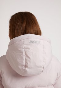 Lichtroze pufferjas met een grote capuchon, voorzien van een subtiel logo op de achterkant en een gladde, gestructureerde stof.