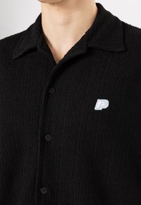 Black textured shirt featuring a spread collar, three buttons, and a small embroidered logo on the chest. Ribbed fabric adds depth.