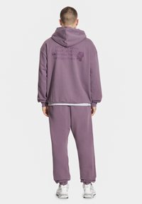 Fioletowa bluza i pasujące spodnie z miękkiego materiału. Bluza ma haftowany tekst i luźny krój. Obuwie to jasne buty sportowe.