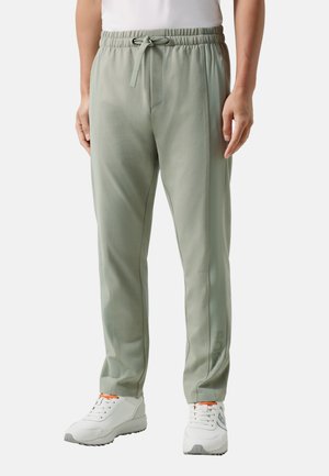 Mand iført lysegrønne bukser med snore og hvide sneakers med orange accenter, stående mod en ensartet baggrund.