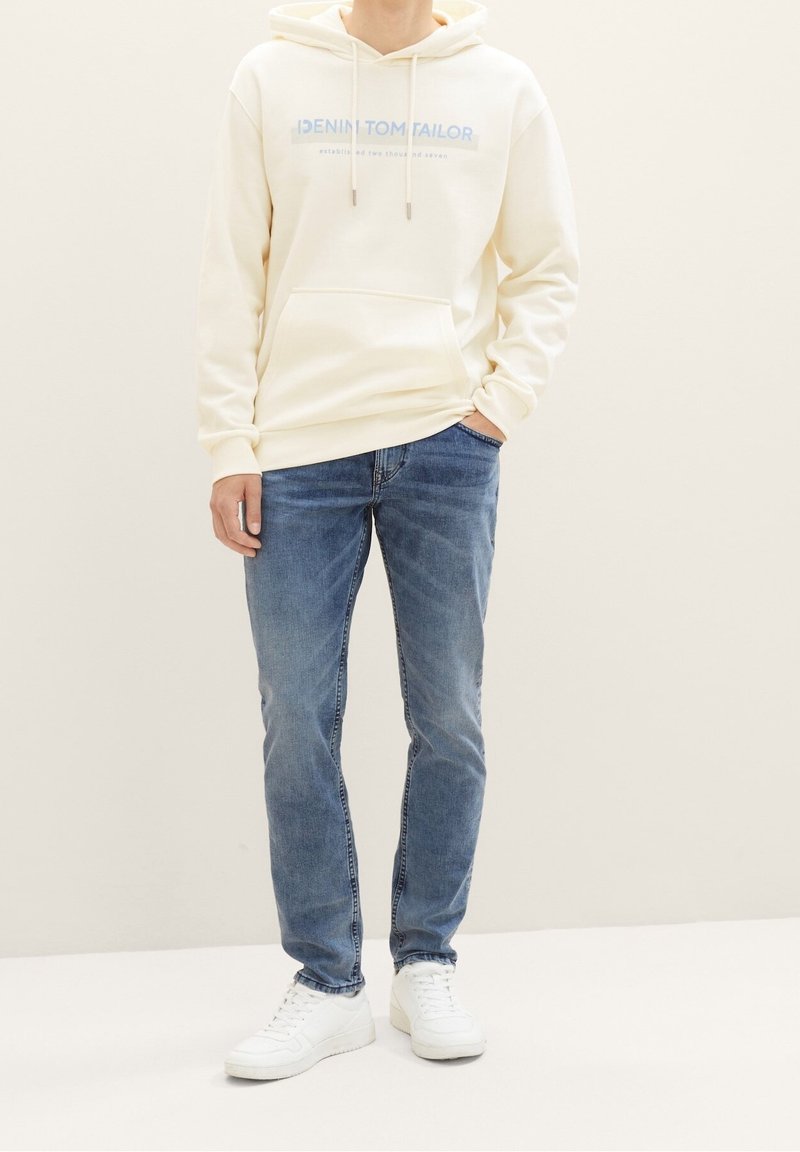 Personne portant un sweat à capuche crème avec un texte bleu clair, un jean bleu, des baskets blanches, debout avec une main dans la poche devant un fond uni.
