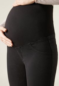 Svarta gravidjeans med hög midja, med en mjuk textur och sidfickor. Händerna vilar på en rundad mage.