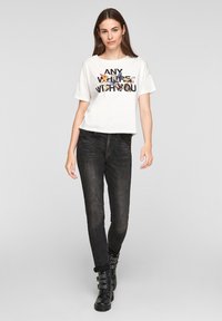 Jeune femme portant un t-shirt graphique blanc avec des papillons et un jean skinny noir, marchant avec assurance dans des bottes de cheville noires à clous.