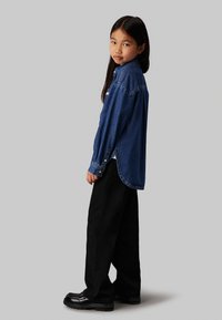 Camicia di jeans blu scuro con colletto a bottoni, maniche lunghe e orlo curvo. Abbinata a pantaloni neri a gamba larga e scarpe nere lucide.