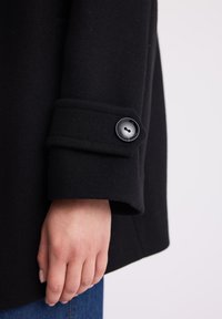 Czarny wełniany płaszcz z podwiniętym mankietem, ozdobiony czarnym guzikiem na rękawie. Gładka faktura i wyrazista konstrukcja.