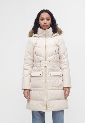 Mujer con abrigo de invierno acolchado beige con capucha forrada de piel, cintura con cinturón, bolsillos delanteros y cremallera dorada, de pie frente a un fondo liso.