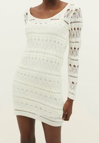 Robe blanche en crochet à manches longues, avec un motif texturé, un design ajouré et une silhouette ajustée. Encolure dégagée et ourlet côtelé.