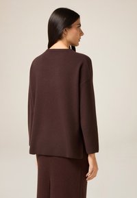 Brauner Strickpullover, lockere Passform, runder Ausschnitt, lange Ärmel, strukturierte Oberfläche. Modell von hinten gezeigt, kombiniert mit passendem Rock.