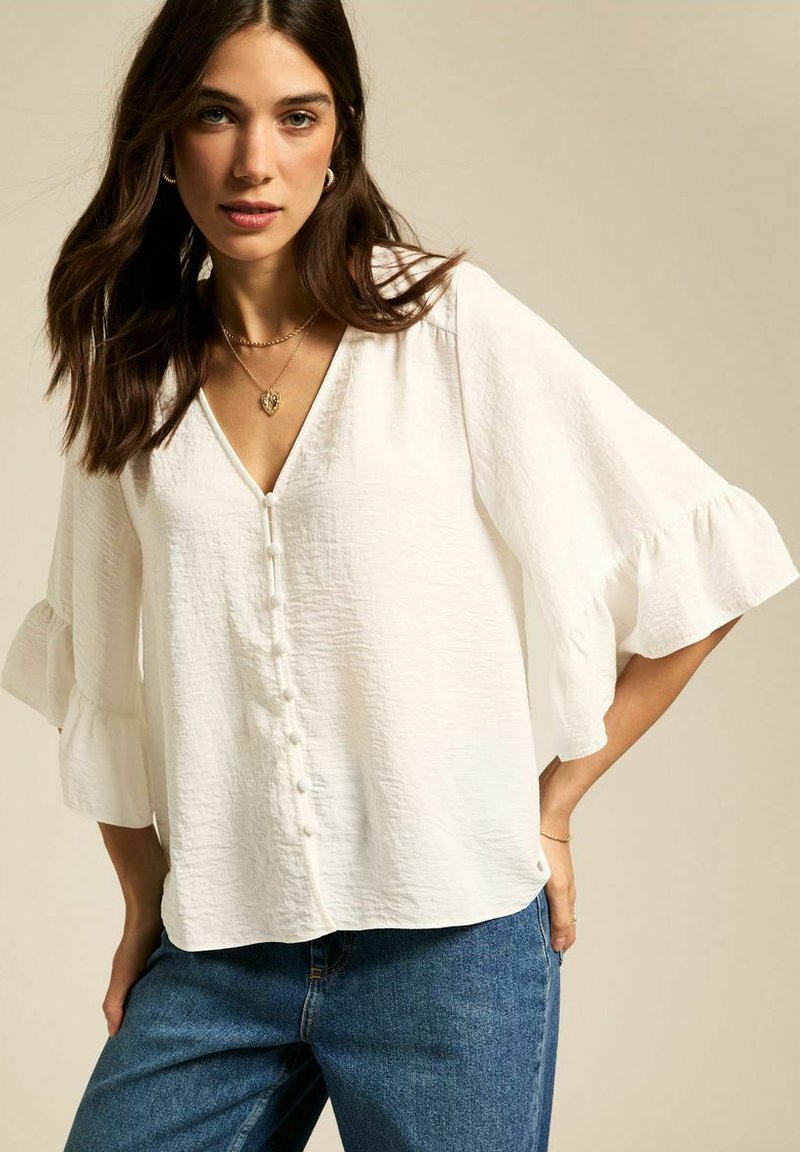 Witte textuur blouse met een V-hals, knoopsluiting en uitlopende driekwart mouwen. Gepaard met blauwe denim jeans.