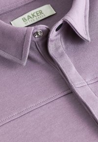 Šviesiai purpurinė polo marškinėliai su apykakle, sidabriniu mygtuku ir subtiliais siuvinėjimo detalėmis. Etiketėje užrašyta "BAKER by Ted Baker."