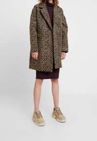 Luipaardprint jas met een losse pasvorm, lange mouwen en een geknipte kraag, gecombineerd met een donkerbruine jurk en chunky beige sneakers.