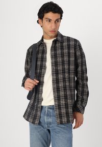 Checkered flannel shirt in black and beige, button-up style, with long sleeves and a relaxed fit. Paired with light blue denim jeans.