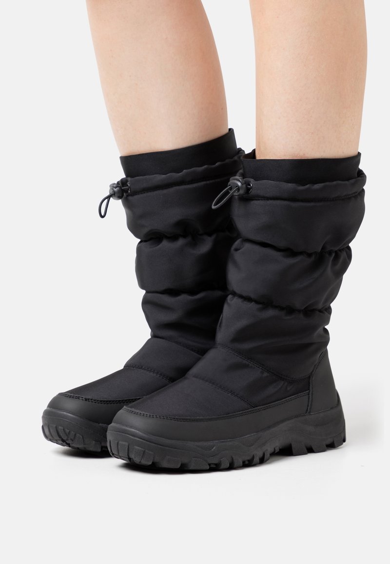 Paire de bottes d'hiver noires isolées avec des guêtres réglables à cordon, portées sur les jambes inférieures d'une personne sur un fond uni.