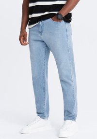 Jeans de mezclilla azul claro con corte slim, que presentan un diseño clásico de cinco bolsillos y detalles de costura, combinados con zapatillas blancas.