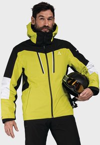 Giacca da sci, di un vivace verde lime con accenti neri e bianchi, tessuto impermeabile, colletto alto, chiusura frontale con zip, design a contrasto, indossata con un casco.