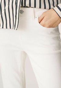 Bílé džíny z denimu s obtaženým střihem, které mají přední kapsu a stříbrné knoflíkové zapínání. Textura vypadá hladce a mírně elasticky.