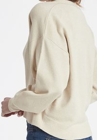 Beige neule, jossa on löysä istuvuus, kammottava helma ja pitkät hihat. Valmistettu teksturoidusta neuleesta, jossa on olkalinja-yksityiskohtia.