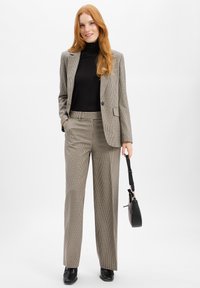 Frau mit roten Haaren, die einen karierten Anzug und einen schwarzen Rollkragenpullover trägt, ein schwarzes Handtasche in einer Hand hält und vor einem weißen Hintergrund lächelt.